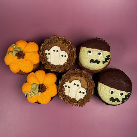 Six Halloween cupcakes featuring pumpkins, ghosts, and zombies on a purple background.
